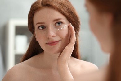 Beautiful woman with freckles applying cream onto her face near mirror in bathroom