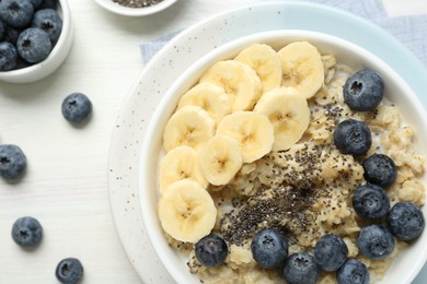 Photo of Tasty oatmeal with banana, blueberries and chia seeds served in bowl on white wooden table, flat lay