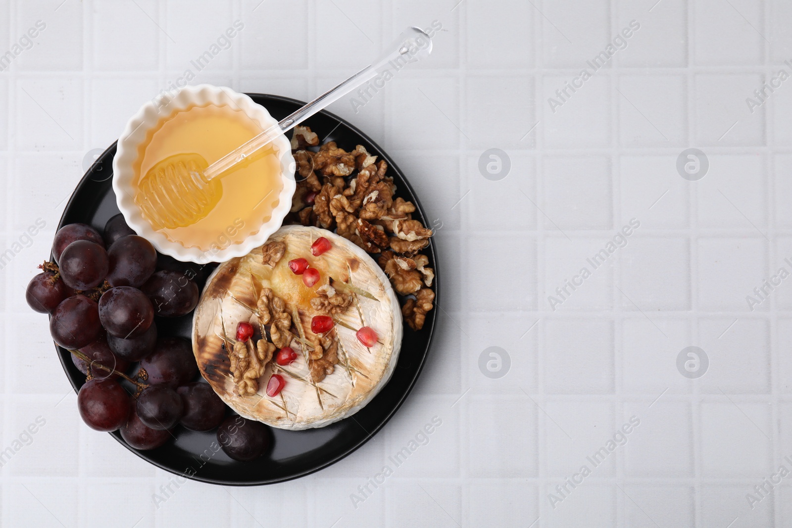 Photo of Plate with tasty baked camembert, honey, grapes, walnuts and pomegranate seeds on white tiled table, top view. Space for text