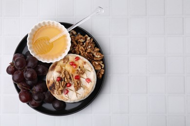 Photo of Plate with tasty baked camembert, honey, grapes, walnuts and pomegranate seeds on white tiled table, top view. Space for text