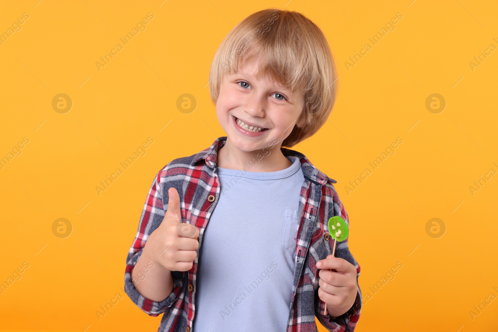Photo of Happy little boy with lollipop showing thumbs up on orange background