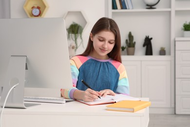 Photo of Cute girl writing in notepad while using computer at desk in room. Home workplace