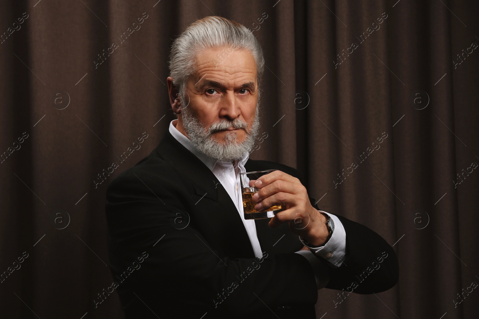 Photo of Senior man in formal suit holding glass of whiskey with ice cubes on brown background