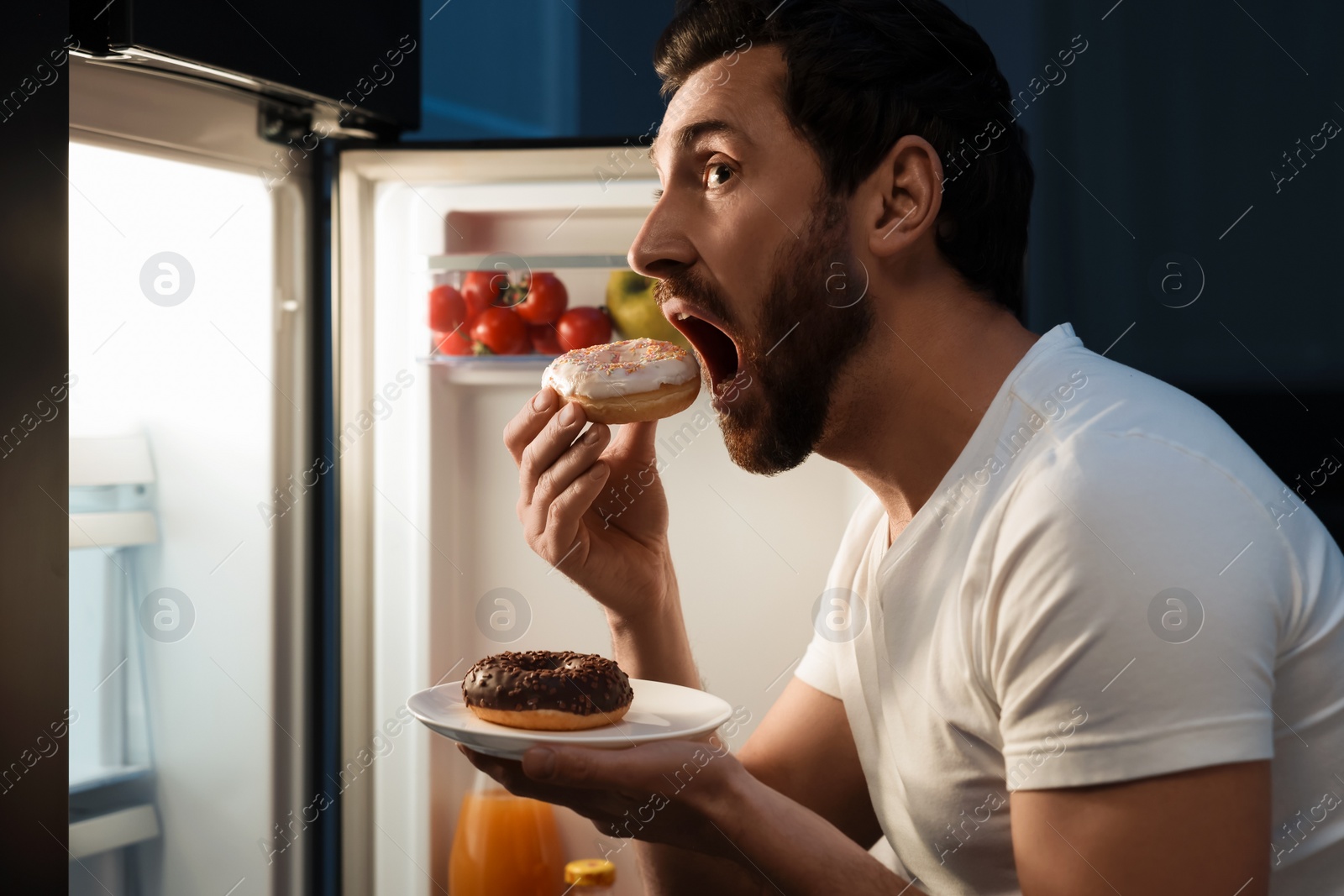 Photo of Man eating donuts near refrigerator in kitchen at night. Bad habit