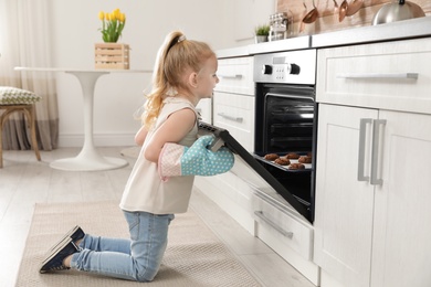 Photo of Little girl opening door of oven with cookies in kitchen