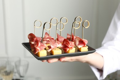 Photo of Woman holding plate of tasty canapes with ham and cheese indoors, closeup
