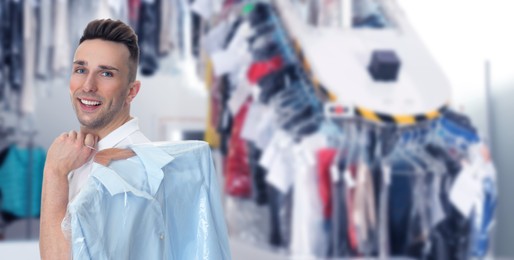 Image of Dry-cleaning service. Happy man holding hanger with shirt in plastic bag indoors, space for text. Banner design