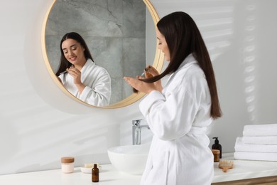 Happy young woman applying essential oil onto hair near mirror in bathroom
