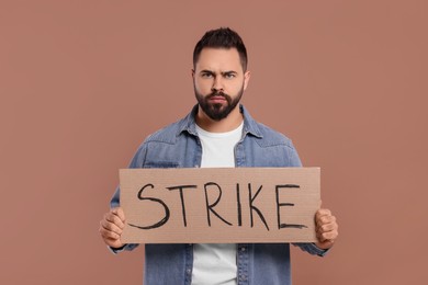 Angry young man holding cardboard banner with word Strike on brown background