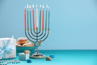 Photo of Hanukkah celebration. Menorah with burning candles, dreidels, donuts and gift boxes on table against light blue background. Space for text
