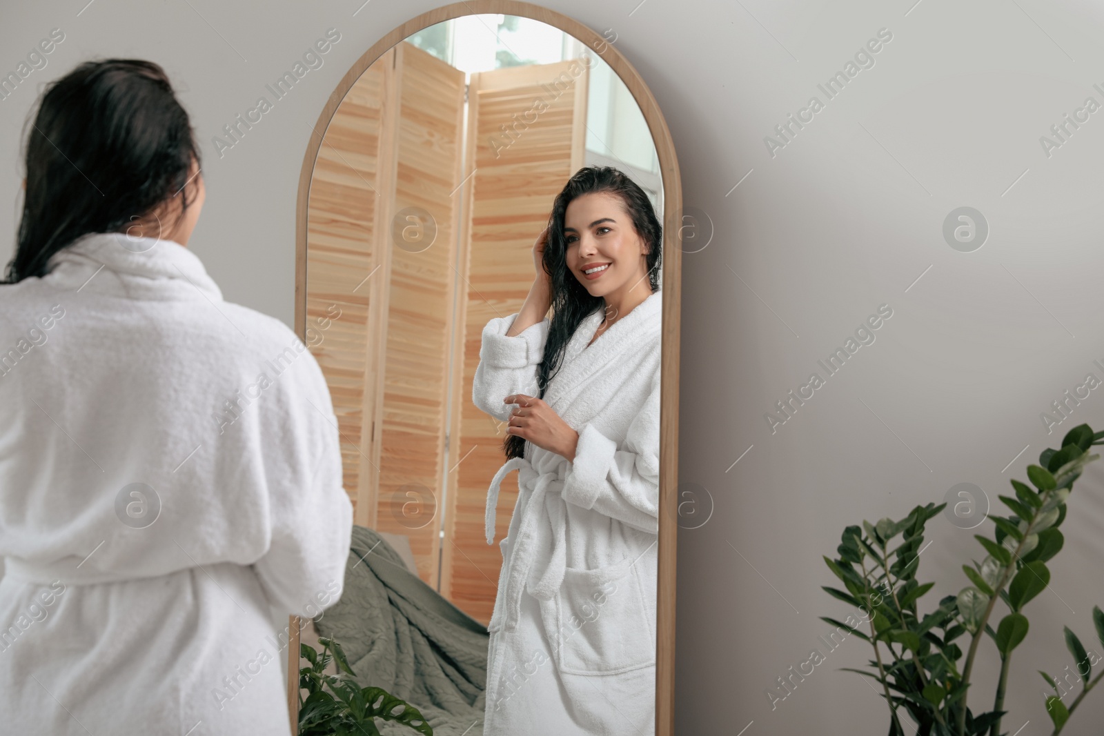 Photo of Beautiful young woman in bathrobe standing near mirror indoors