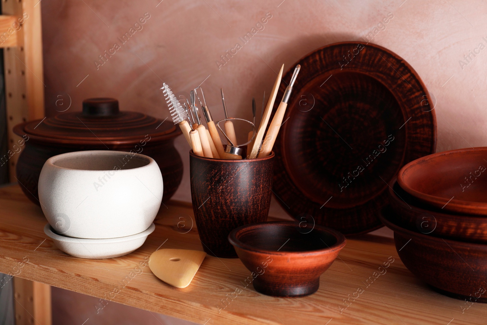 Photo of Set of different crafting tools and clay dishes on wooden rack in workshop
