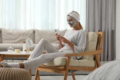 Young woman with face mask using smartphone at home, space for text. Spa treatments