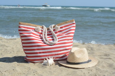 Photo of Stylish striped bag with straw hat and seashell on sandy beach near sea
