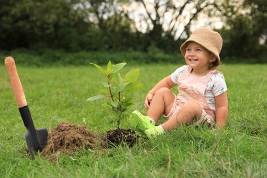 Cute happy baby girl near young green tree and trowel in garden