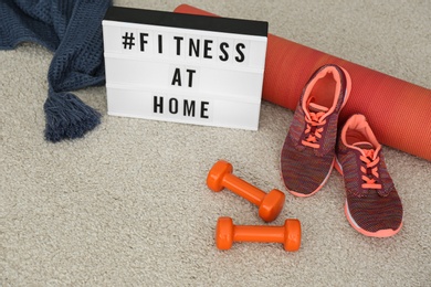 Photo of Sport equipment and lightbox with hashtag FITNESS AT HOME on carpet. Message to promote self-isolation during COVID‑19 pandemic