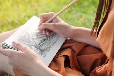 Photo of Young woman drawing with pencil in notepad outdoors on sunny day, closeup