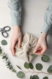 Photo of Furoshiki technique. Woman decorating gift wrapped in fabric with eucalyptus branch at white wooden table, top view