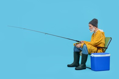 Fisherman with rod and cool box on chair against light blue background