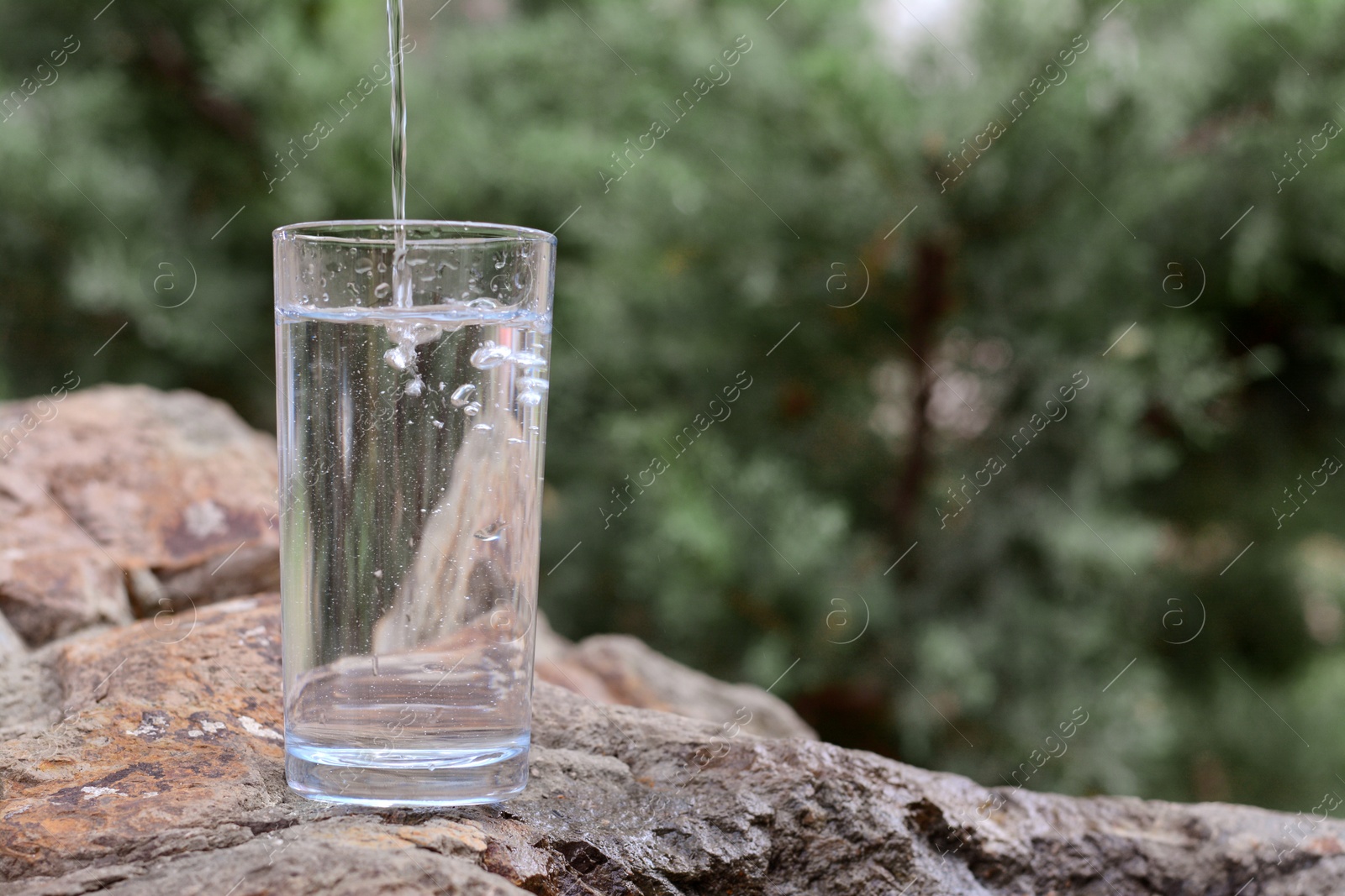 Photo of Pouring pure water into glass on stone outdoors, space for text