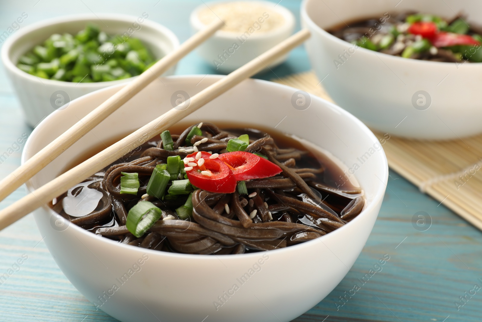 Photo of Tasty soup with buckwheat noodles (soba), chili pepper, green onion in bowl and chopsticks on light blue wooden table, closeup