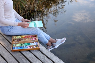 Photo of Woman drawing with soft pastels on wooden pier near river, closeup