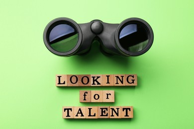 Photo of Staff recruitment concept. Phrase Looking For Talent made of wooden cubes and binoculars on light green background, flat lay