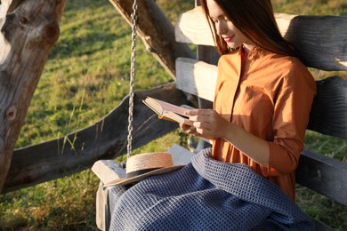 Photo of Beautiful young woman reading book on wooden swing outdoors