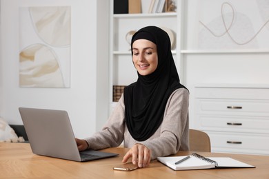 Photo of Muslim woman in hijab using laptop at wooden table in room