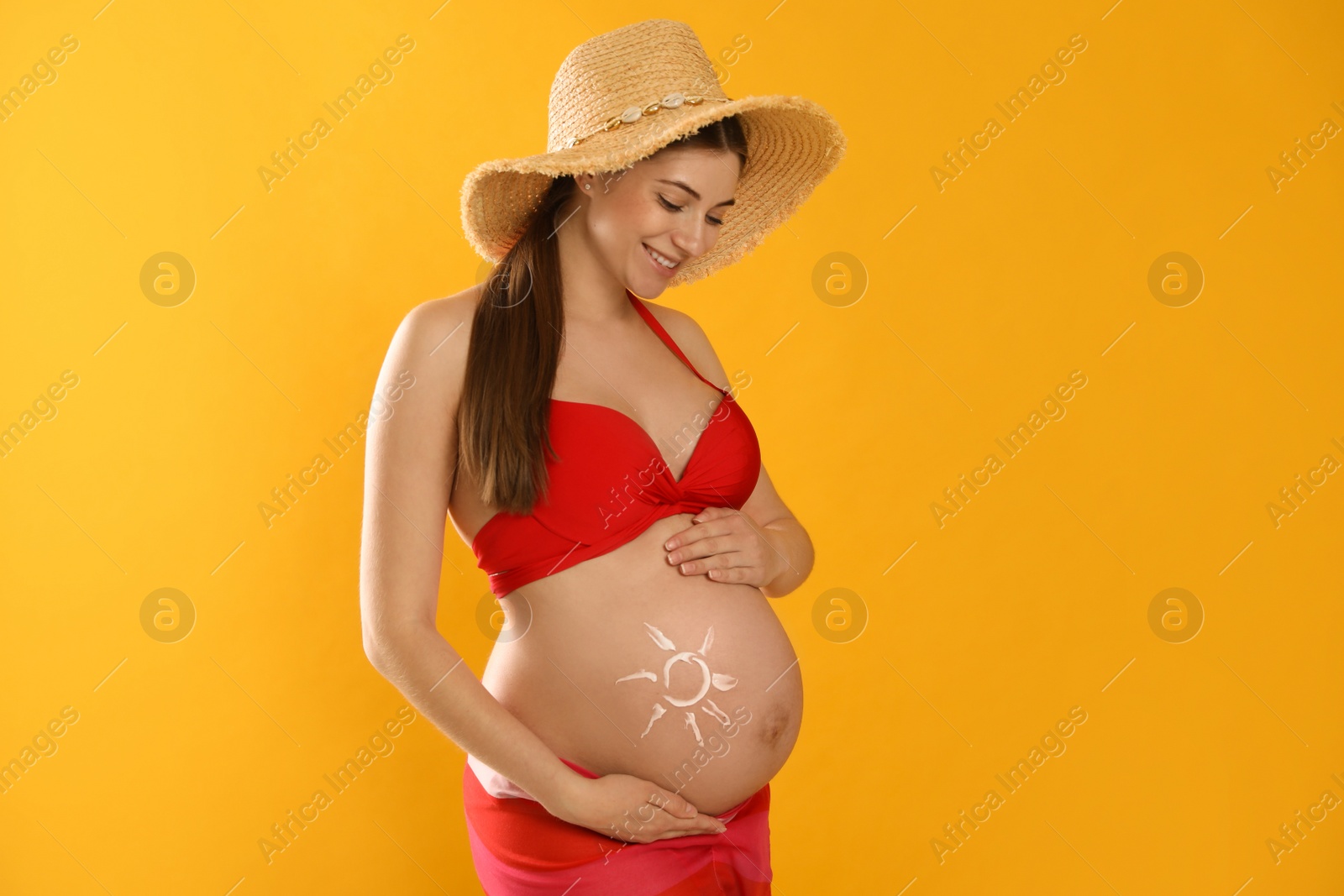 Photo of Young pregnant woman with sun protection cream on belly against yellow background