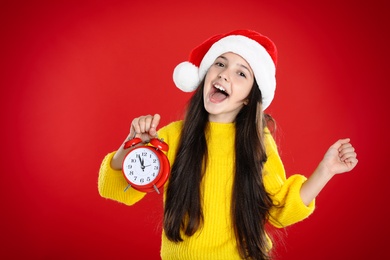 Girl in Santa hat with alarm clock on red background. New Year countdown