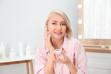 Portrait of charming mature woman with healthy beautiful face skin and natural makeup applying cream indoors