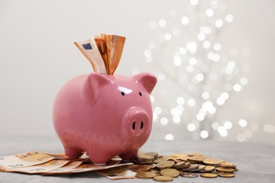 Photo of Piggy bank with euro banknotes and coins on grey table against blurred lights, space for text