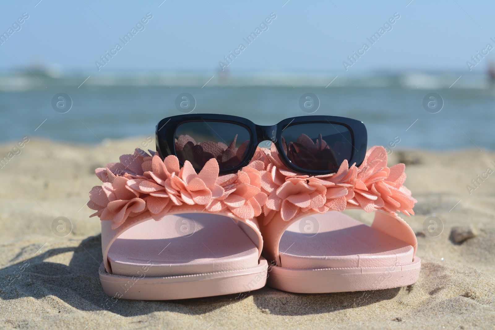 Photo of Stylish slippers and sunglasses on sandy beach near sea, closeup