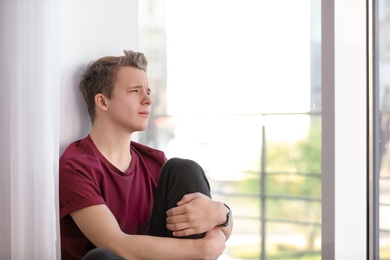 Photo of Upset teenage boy sitting alone near window indoors