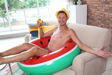 Photo of Shirtless man with inflatable ring and bottle of drink on sofa at home