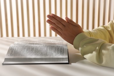 Woman holding hands clasped while praying over Bible at white table indoors, closeup