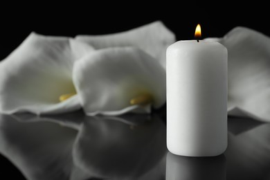 Photo of Burning candle and white calla lily flowers on black mirror surface in darkness, closeup with space for text. Funeral symbol