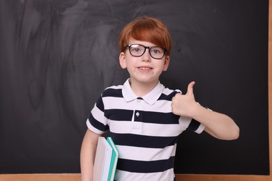 Photo of Smiling schoolboy in glasses with books showing thumb up near blackboard