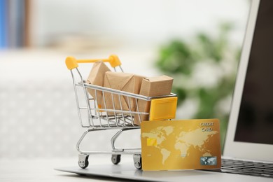 Photo of Online payment concept. Small shopping cart with bank card, boxes and laptop on table