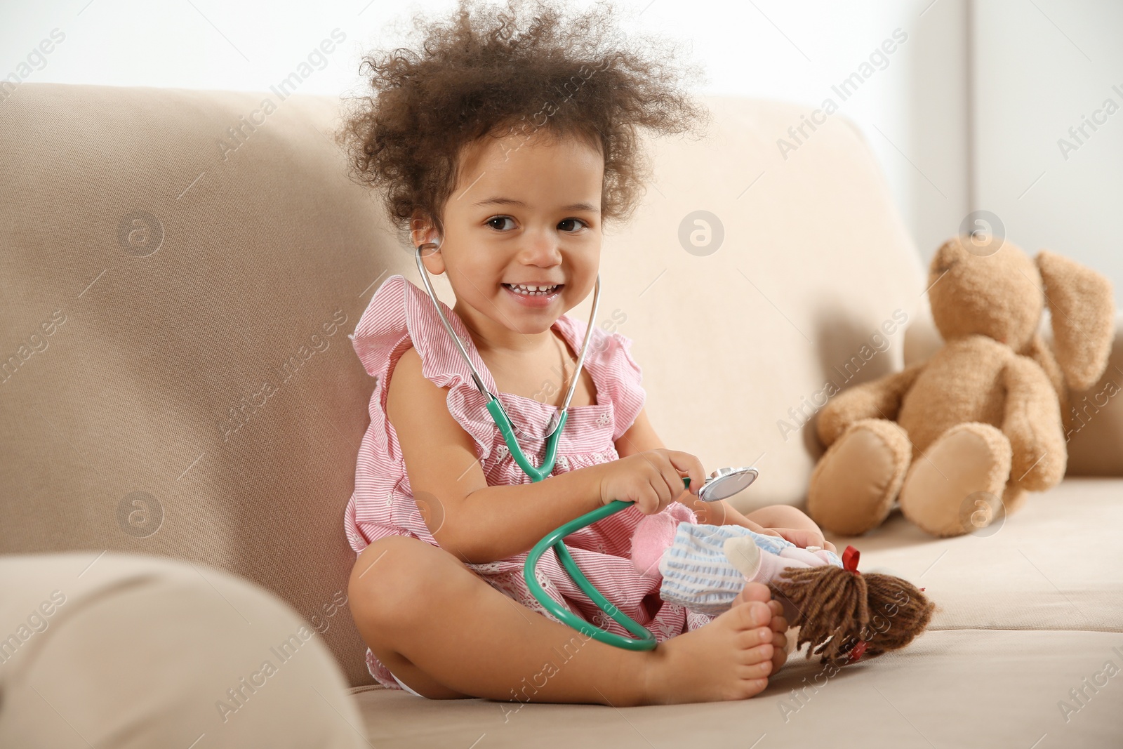 Photo of Cute African American child imagining herself as doctor while playing with stethoscope and doll on couch at home