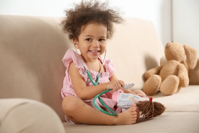 Photo of Cute African American child imagining herself as doctor while playing with stethoscope and doll on couch at home