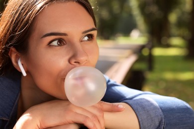 Photo of Beautiful young woman with wireless headphones blowing chewing gum outdoors, closeup