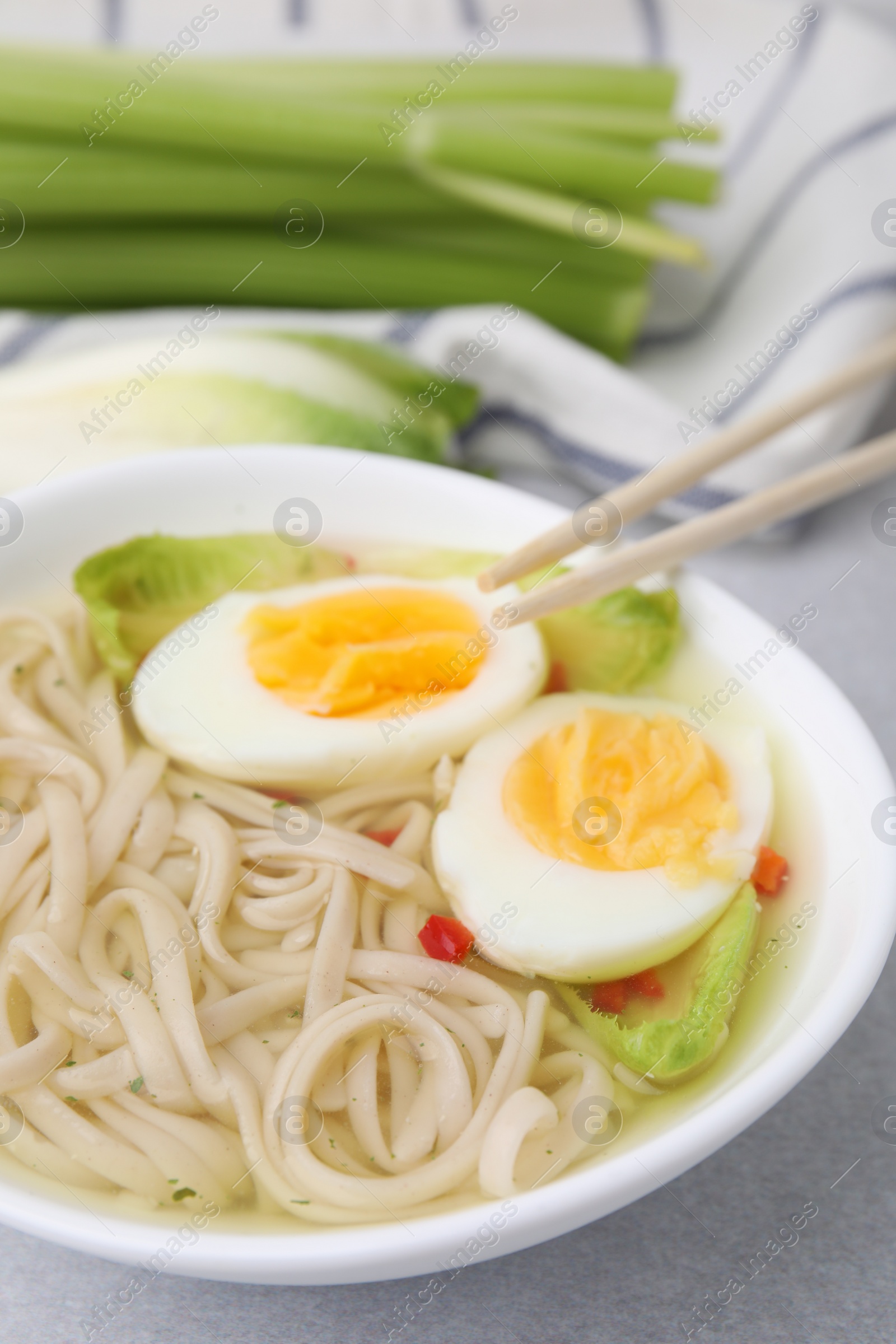 Photo of Bowl of delicious rice noodle soup with celery and egg on light grey table, closeup