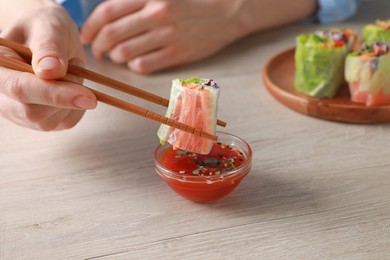 Woman dipping delicious spring roll wrapped in rice paper into sauce at white wooden table, closeup