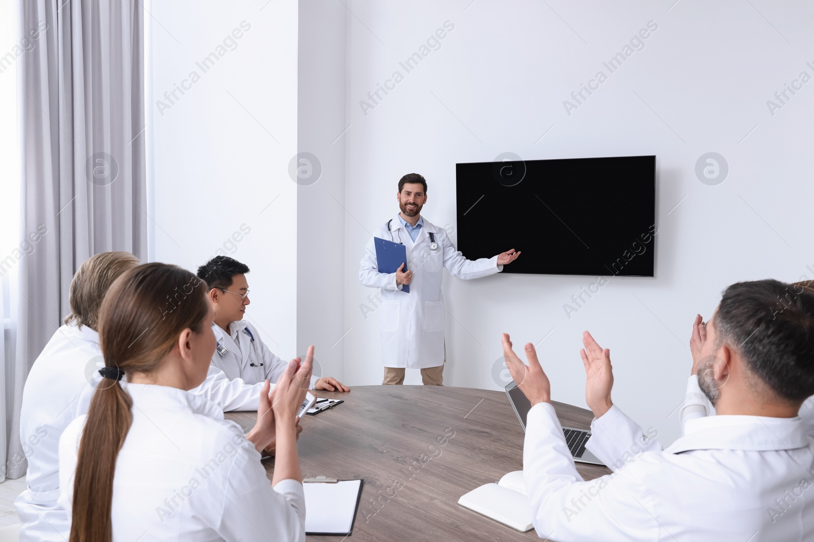 Photo of Team of doctors listening to speaker report near tv screen in meeting room. Medical conference