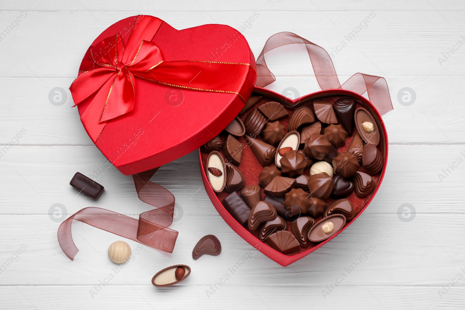 Photo of Heart shaped box with delicious chocolate candies and ribbon on white wooden table, flat lay