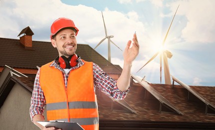 Image of Industrial engineer in uniform and view of wind energy turbines near house with installed solar panels on roof