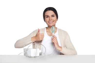 Photo of Happy young woman with nebulizer showing thumb up at table on white background