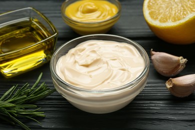 Photo of Glass bowl with fresh mayonnaise and ingredients on black wooden table, closeup